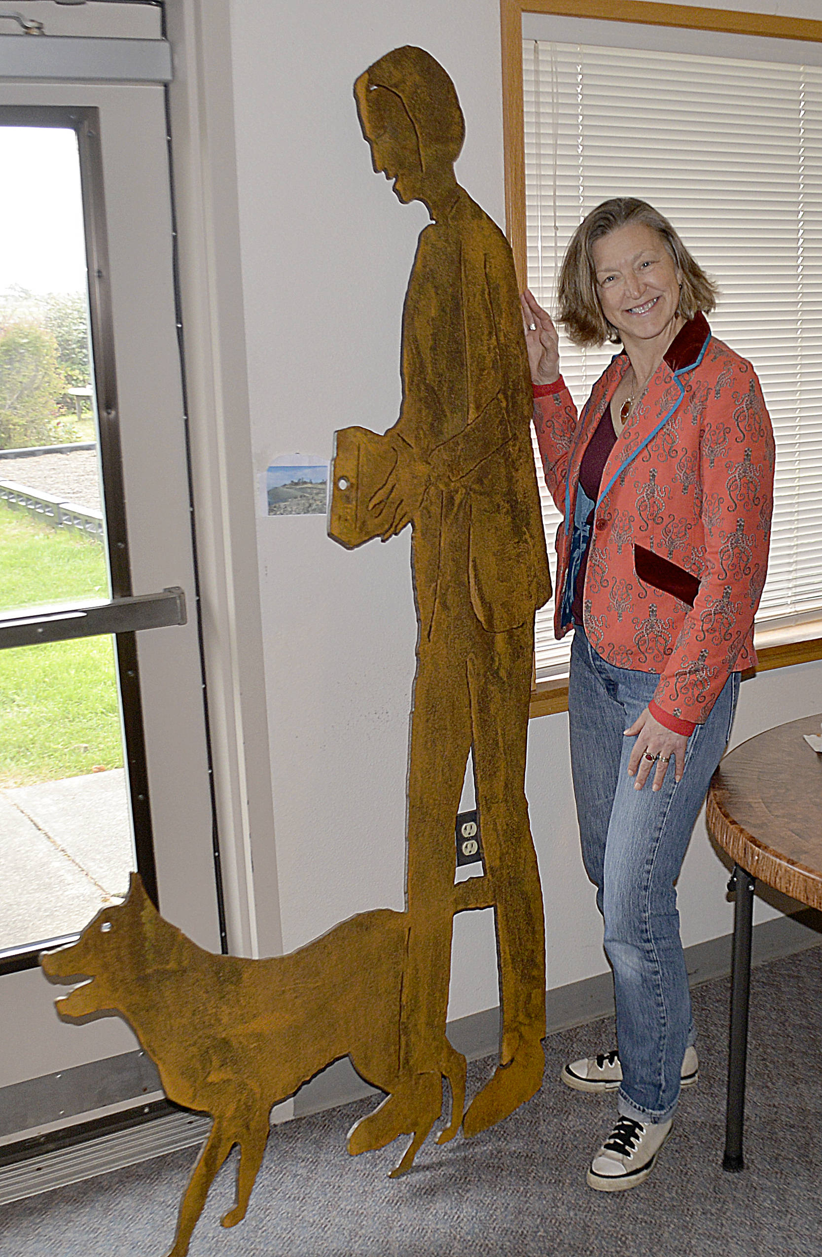 DAN HAMMOCK | GRAYS HARBOR NEWS GROUP                                North Cove resident Marcy Merill poses with a steel cutout she and her dog Hawkeye modeled for, part of the “Shape the Shore” project. The iron silhouette was made by local artist John Jones and will be placed at the revetment project at the end of Old State Route 105. The box camera she is holding in the piece includes a hole that will point visiting photographers to a specific spot in the revetment to take a picture. Those photos can be used to help track the movement of the rock in the revetment.