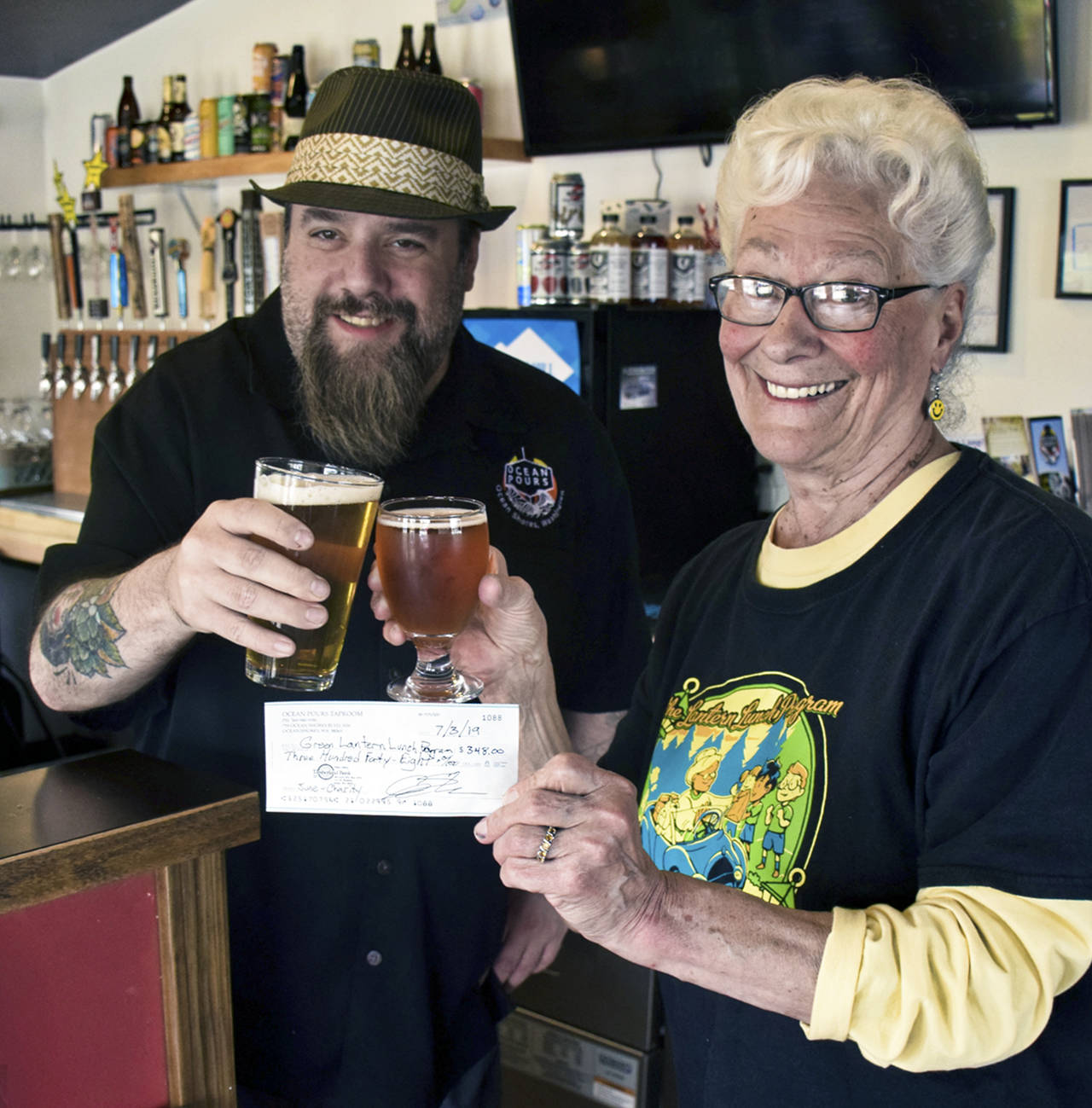 Scott D. Johnston | For Grays Harbor News Group                                Ocean Pours Taproom general manager Chris Shifman, left, and Green Lantern Lunch Program founder Phyllis Shaughnessy toast the pub’s donation to the North Coast nonprofit organization, which feeds area kids during the summer. Shifman says each month, his Ocean Shores pub designates one of more of its 16 taps to give a 50-cent donation for each glass poured to a different local nonprofit. June’s donation for Green Lantern amounted to nearly $350, which he says was the largest amount since the business opened last winter.
