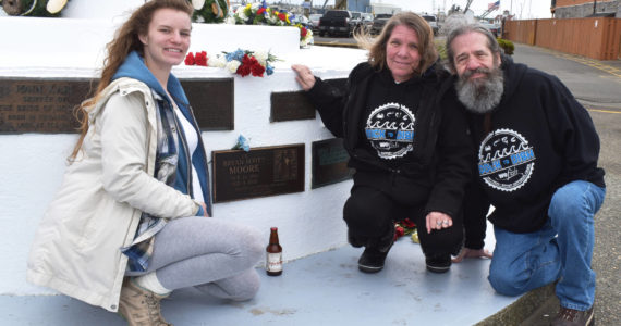 Bryan Scott Moore’s family — his sister Caitlin, and his parents Audrey and John Moore — lay a single blue flower and a Lagunitas Brewing’s “A Little Sumpin Sumpin Ale” at his memorial on Sunday, May 29, 2022, after “The Blessing of the Fleet” at Fisherman’s Memorial in Westport. “Cold beer was pretty much his favorite,” Bryan’s father said. (Matthew N. Wells | The Daily World)