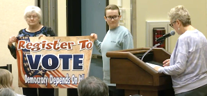 Indivisible Ocean Shores member Maria Van Horn speaks at the March 25 Ocean Shores City Council meeting while supporters hold a politically charged banner. (North Beach TV screenshot)