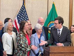 Jerry Cornfield / Washington State Standard
Gov. Bob Ferguson, at podium, goes to shake hands with state Sen. Noel Frame, D-Seattle, at the signing of a bill to make clergy mandatory reporters of child abuse and neglect, on May 2, in Olympia. Standing between them is Mary Dispenza, a founding member of the Catholic Accountability Project.