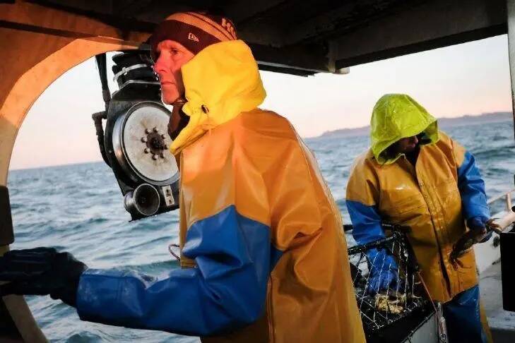 Andy Mitby nudges the steering lever of the fishing vessel Ragnarok while Eric Hopfer measures and sorts crab.