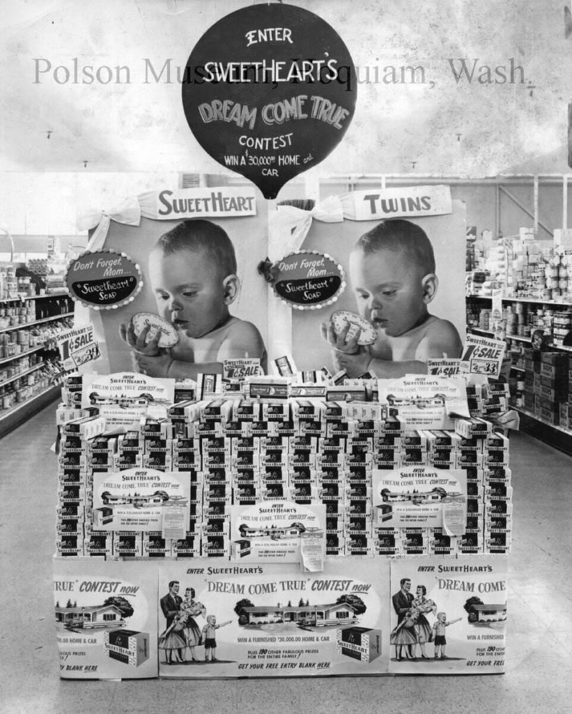 Mark Swanson / Jones Photo Collection
This image shows multiple contest posters around a supermarket display of Sweethearts Soap, which is the contest sponsor. From the top: a balloon-shaped sign has colored lettering on a dark background, pleading Enter Sweethearts Dream Come True Contest, Win a $30,000 Home and Car. This display was evidently at Swansons, probably the Hoquiam store but possibly in Aberdeen. Below that sign are placed two large posters of identical infants appreciatively staring at bars of soap in their hands. Below the twins are stacked an estimated 1,900 boxes of soap. Lastly, more posters wrap the bottom of the display, showing a family of five contemplating a ranch-style home with a station wagon parked in the garage. The automobile pictured resembles a circa 1954 General Motors product.